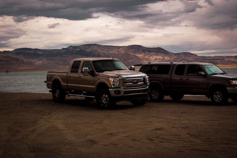 Pickup trucks parked by a serene mountain lake under a cloudy sky, creating a rugged, adventurous scene