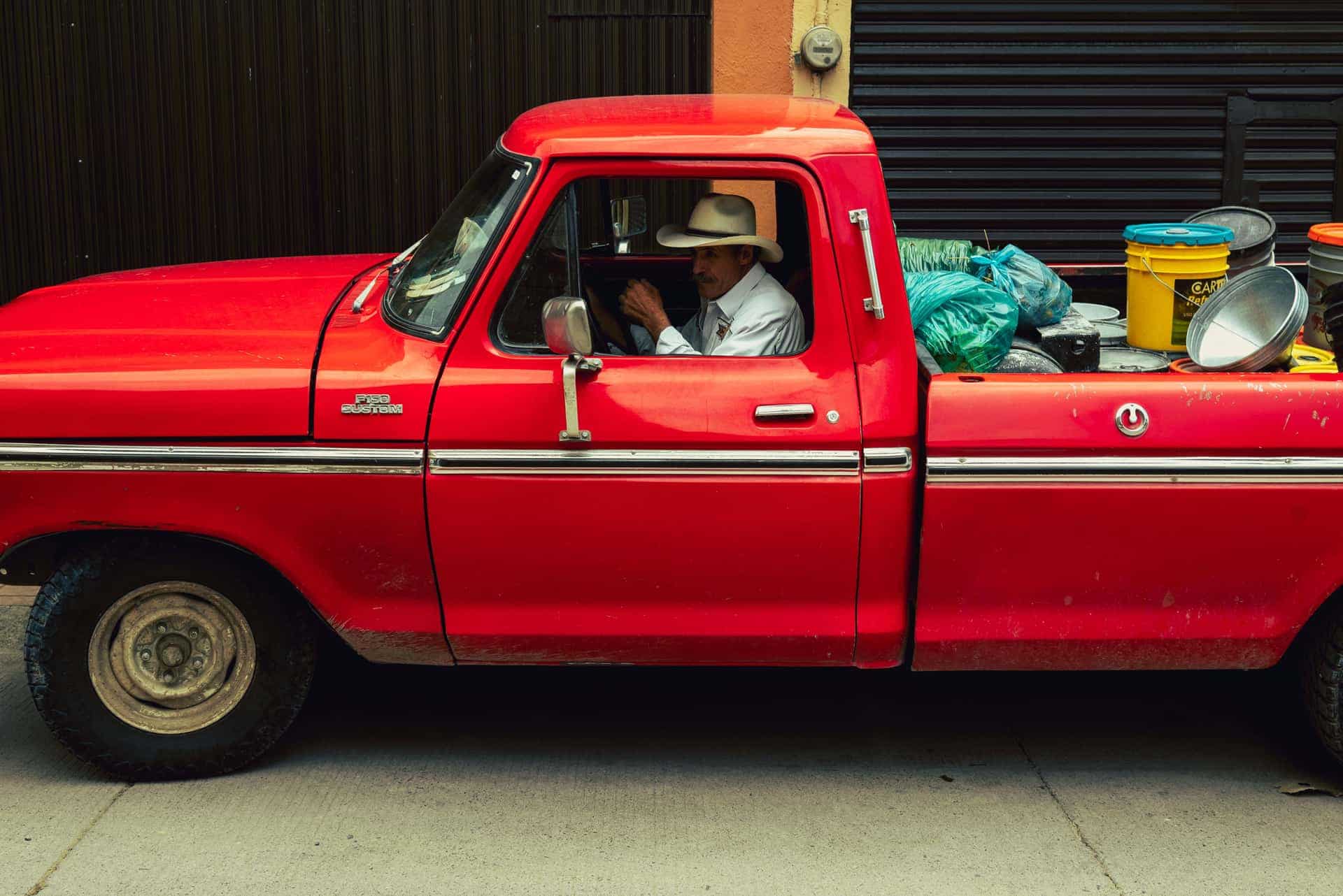 A classic red truck with a driver wearing a hat, parked in an urban area with colorful containers