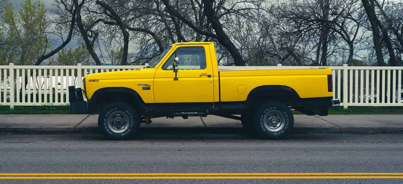 Side view of a parked yellow pickup truck on a city street with trees and a fence in the background