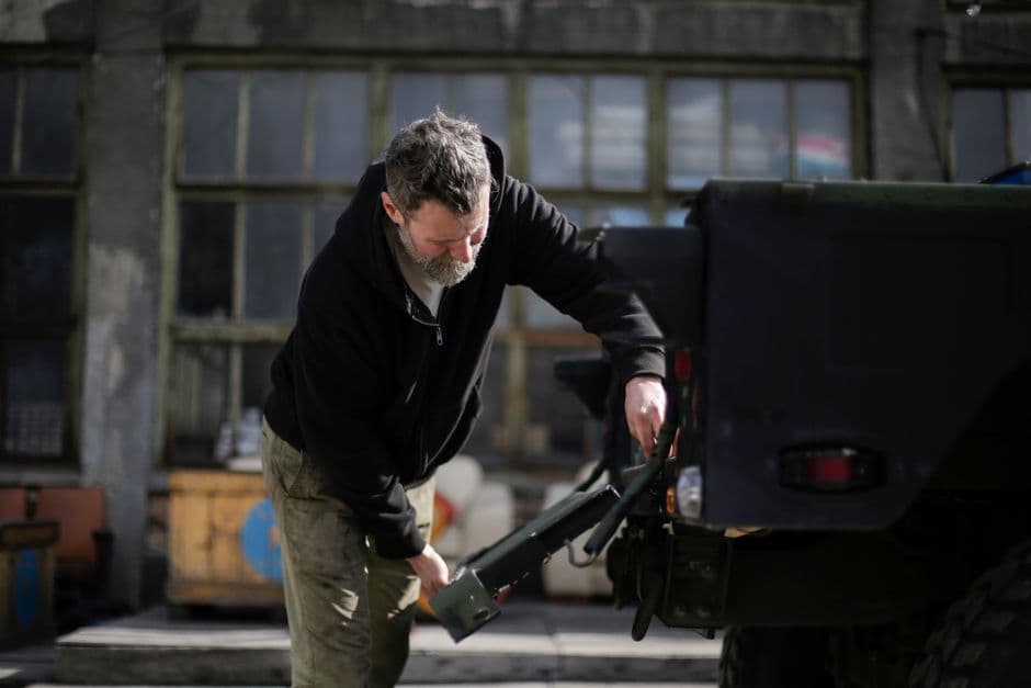 Side view of a bearded mechanic in workwear repairing the metal trunk door of a vehicle on the street near a garage