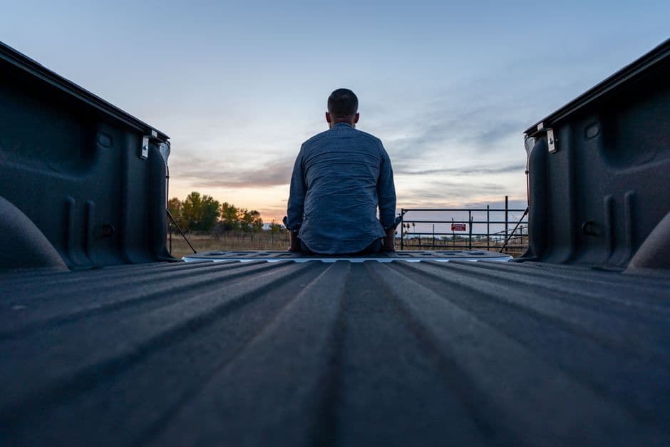 A person sits in a truck bed, facing a scenic sunset outdoors, captured from behind