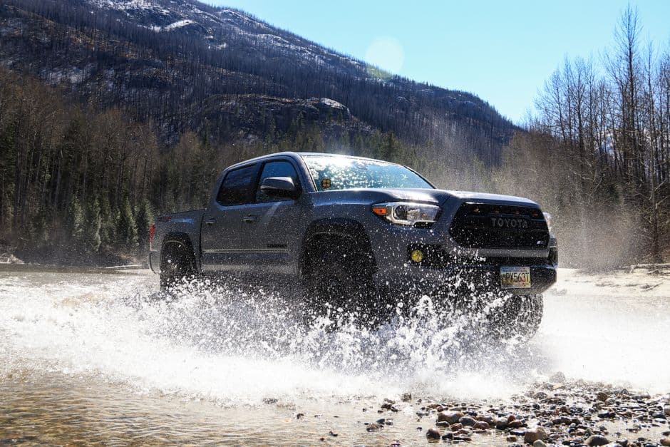 A Toyota Tacoma drives through a water puddle with splashing effects in a rugged outdoor setting