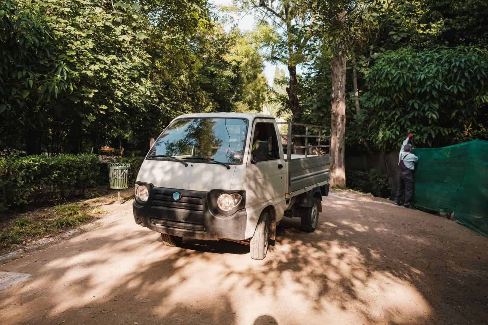 Small white truck parked on a sunny day in a green park with trees and a man working