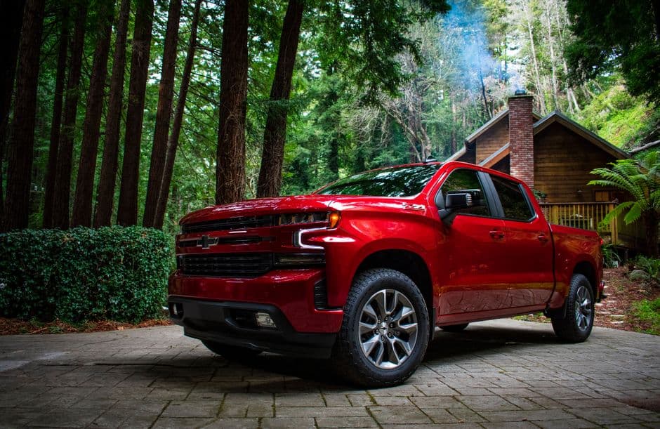 A red Chevrolet Silverado 1500 parked in a rustic driveway surrounded by lush forest and cottage