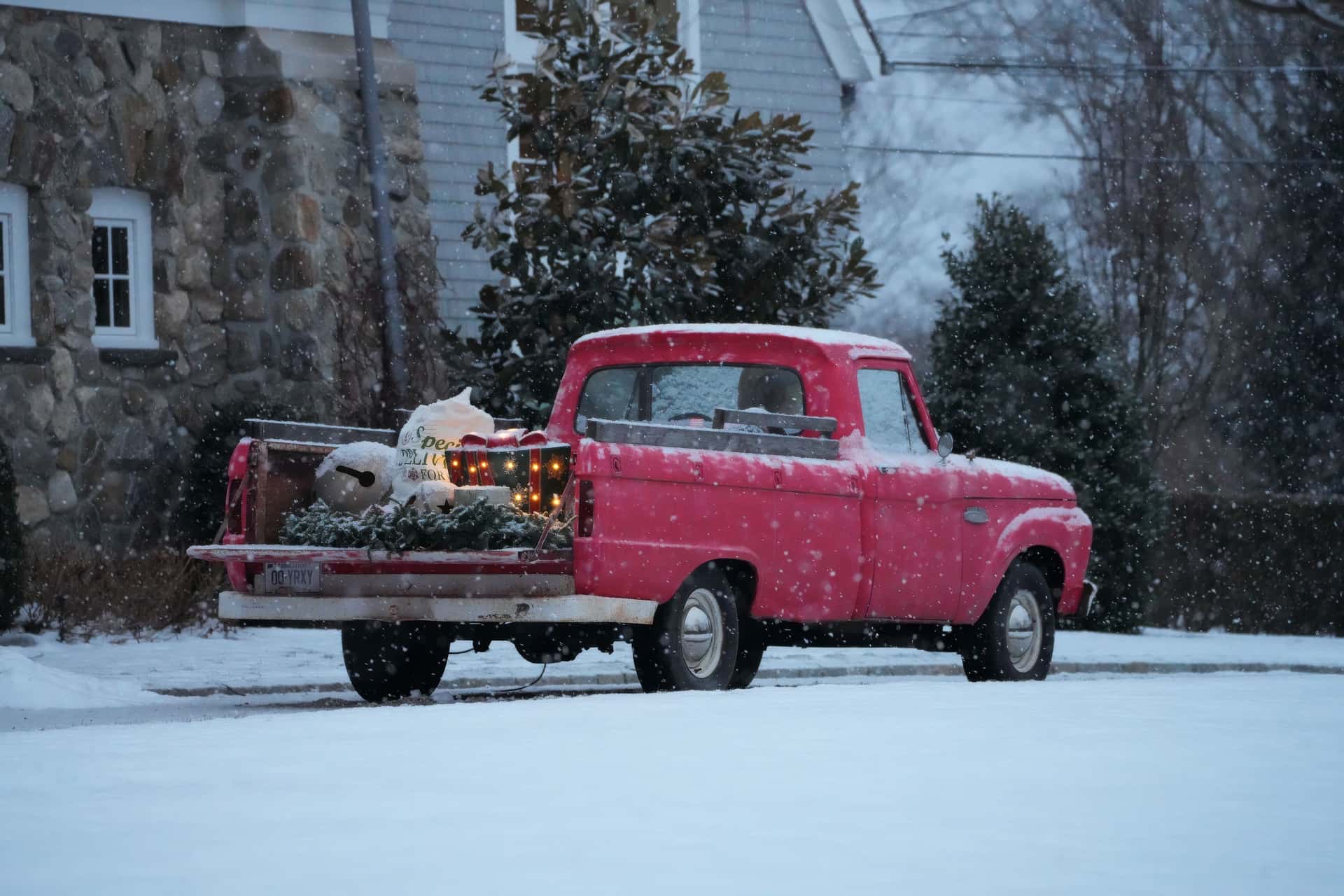 A classic red truck parked in snow, decorated for Christmas with trees and lights